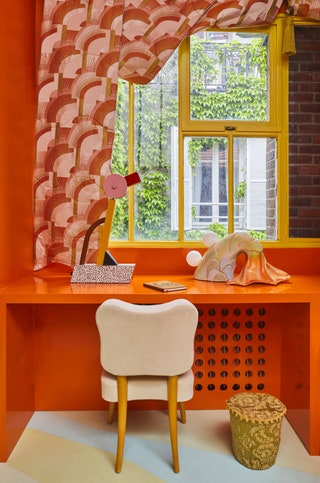 white chair pulled up to orange desk built into orange wall patterned orange drapes around a yellow window show ivy...